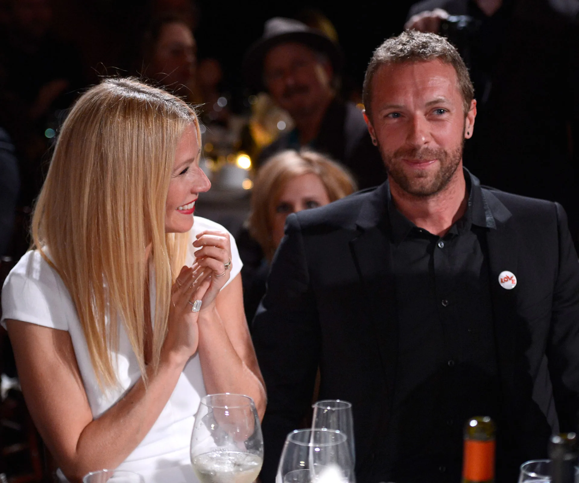 A woman in white smiles and claps next to a man in black at a formal event with wine glasses on the table.