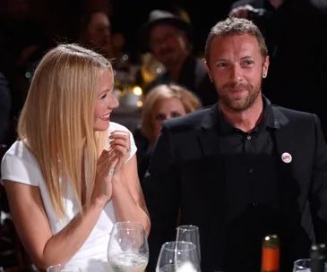 A woman in white smiles and claps next to a man in black at a formal event with wine glasses on the table.