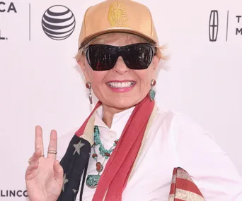 ```plaintext
Person smiling in sunglasses and cap, wearing jewelry and a scarf with flag pattern, against a backdrop with logos.
```