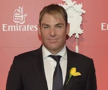A man in a black suit with a yellow flower on the lapel stands against a red Emirates-branded background.