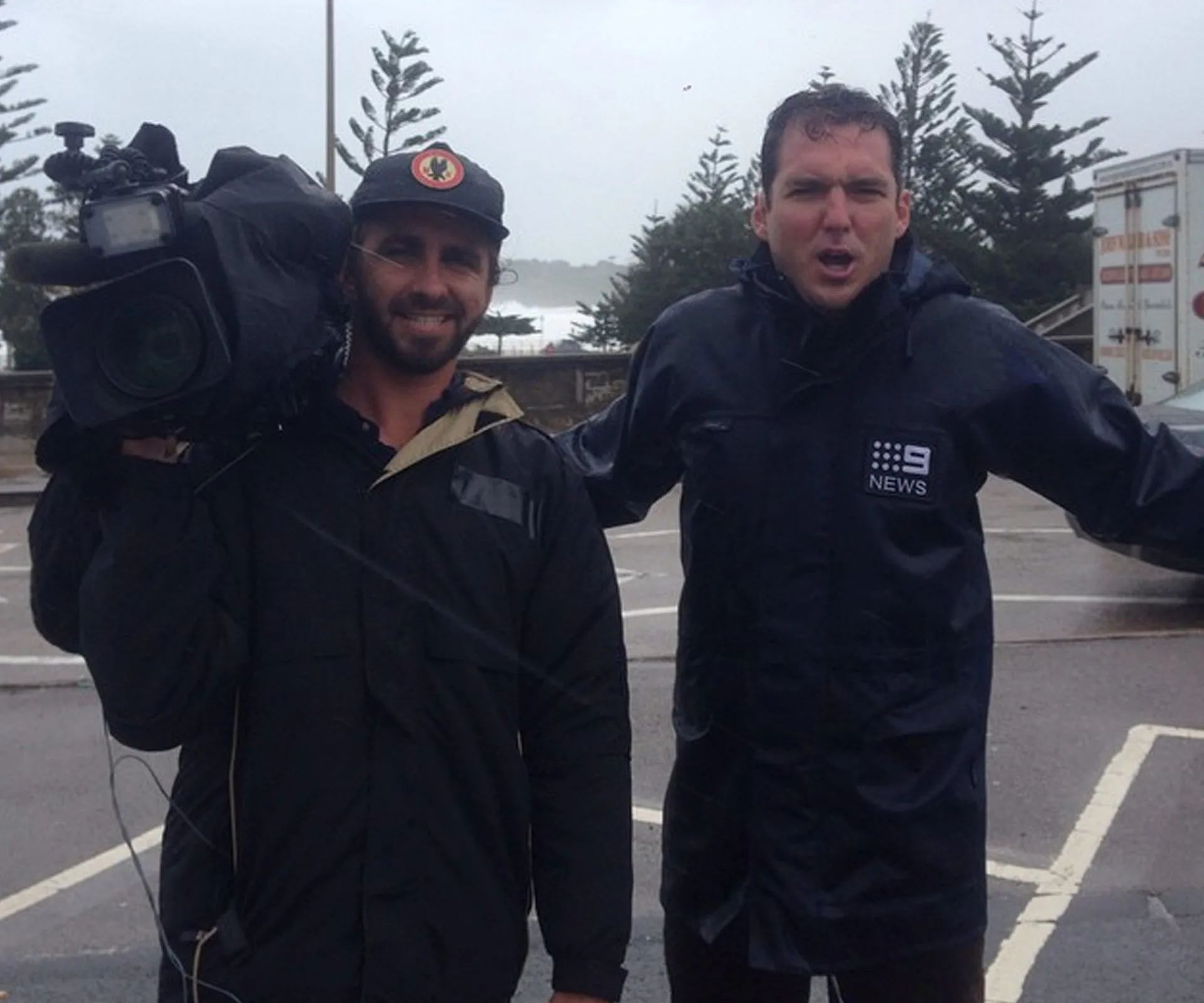 News reporter and cameraman in rain gear braving a storm in a parking lot with trees and vehicles in the background.
