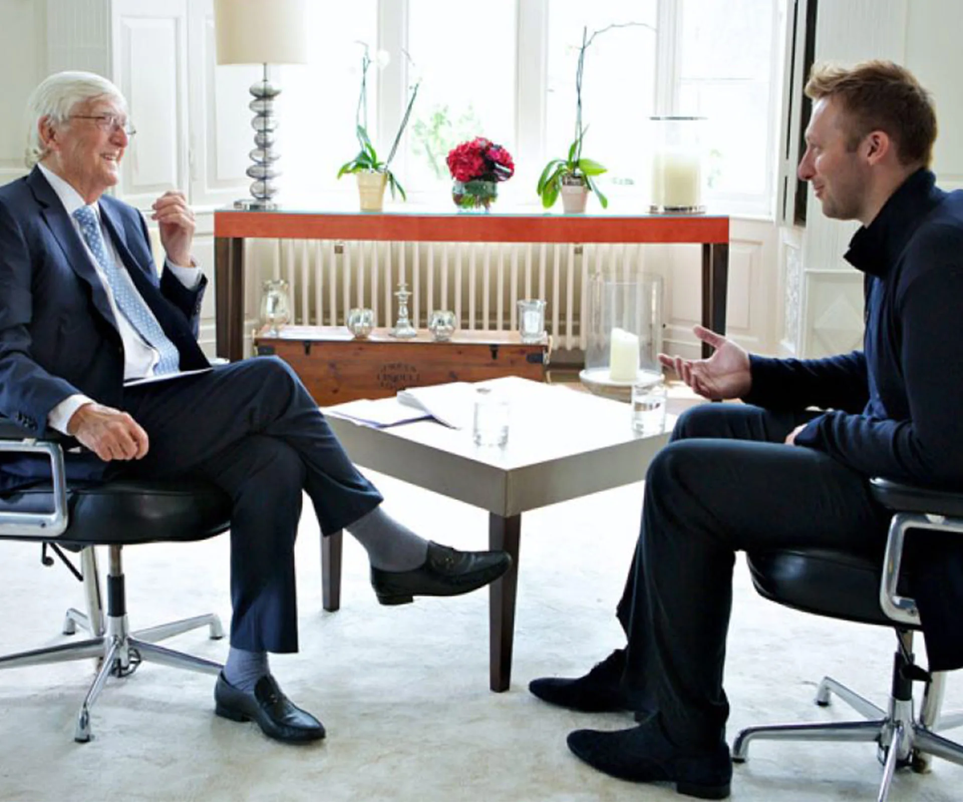 Two men seated and having a conversation in a bright, stylish room with flowers and a table with papers.