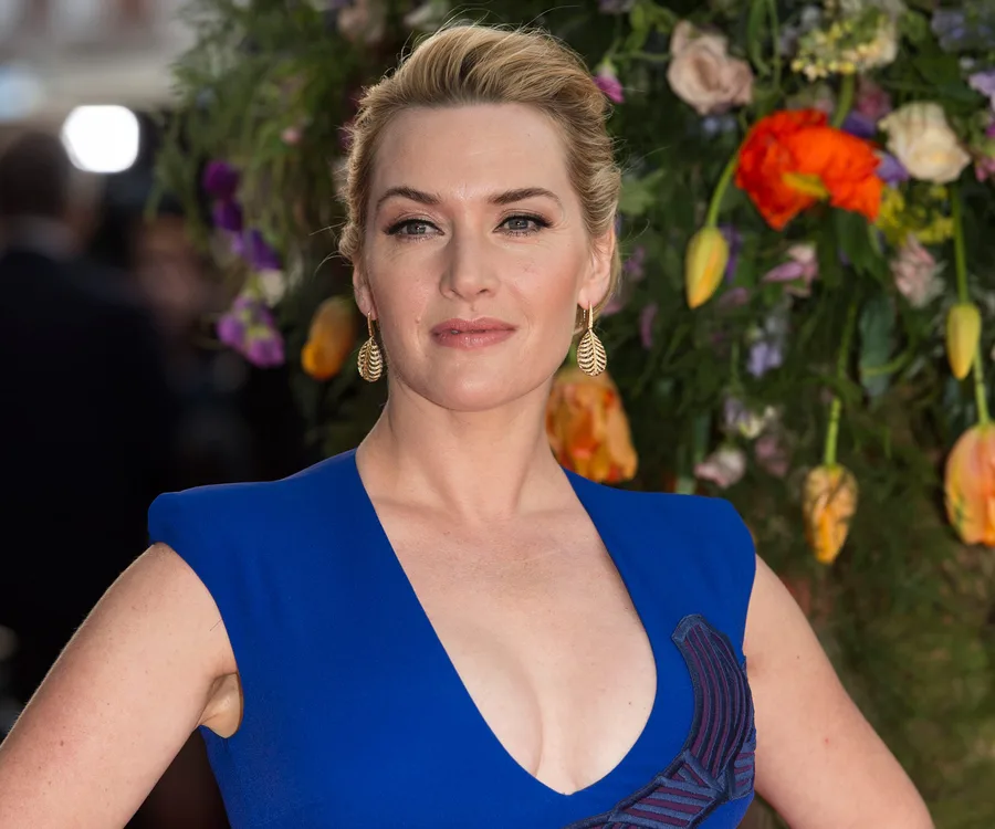 Kate Winslet in a blue dress at a floral backdrop event, wearing leaf-shaped earrings and looking towards the camera.