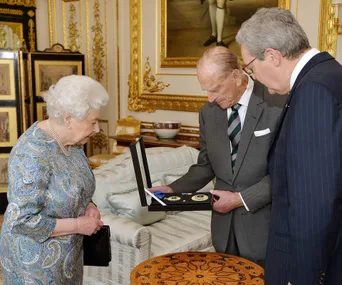 Senior man shows others a medal set in a royal-like room with ornate decor and a gold-framed painting on the wall.