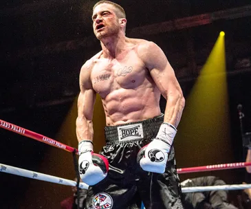 A muscular boxer with tattoos stands in a boxing ring, wearing boxing gloves and shorts, lit by dramatic yellow lighting.