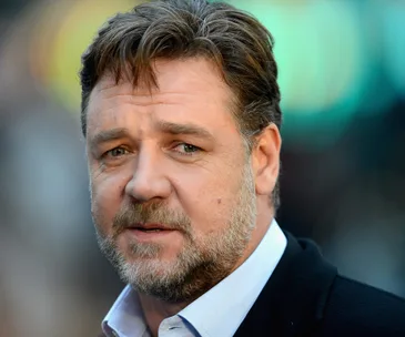 Russell Crowe with a thoughtful expression, wearing a suit, with a blurred background.