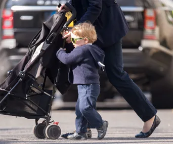 Young child with sunglasses pushes a stroller assisted by an adult in a city street.