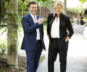 Man and woman in suits stand smiling outdoors next to a tree, appearing cheerful and relaxed.