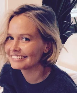 Smiling woman with short blonde hair, makeup-free, in a cozy setting.