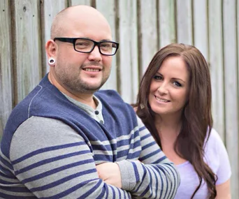 Couple posing in front of a wooden fence, both smiling, the man wearing glasses and striped sweater, the woman in a white top.