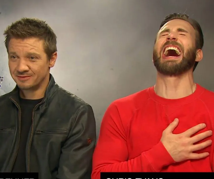 Two men seated, one in a red shirt laughing, the other in a leather jacket with a neutral expression.
