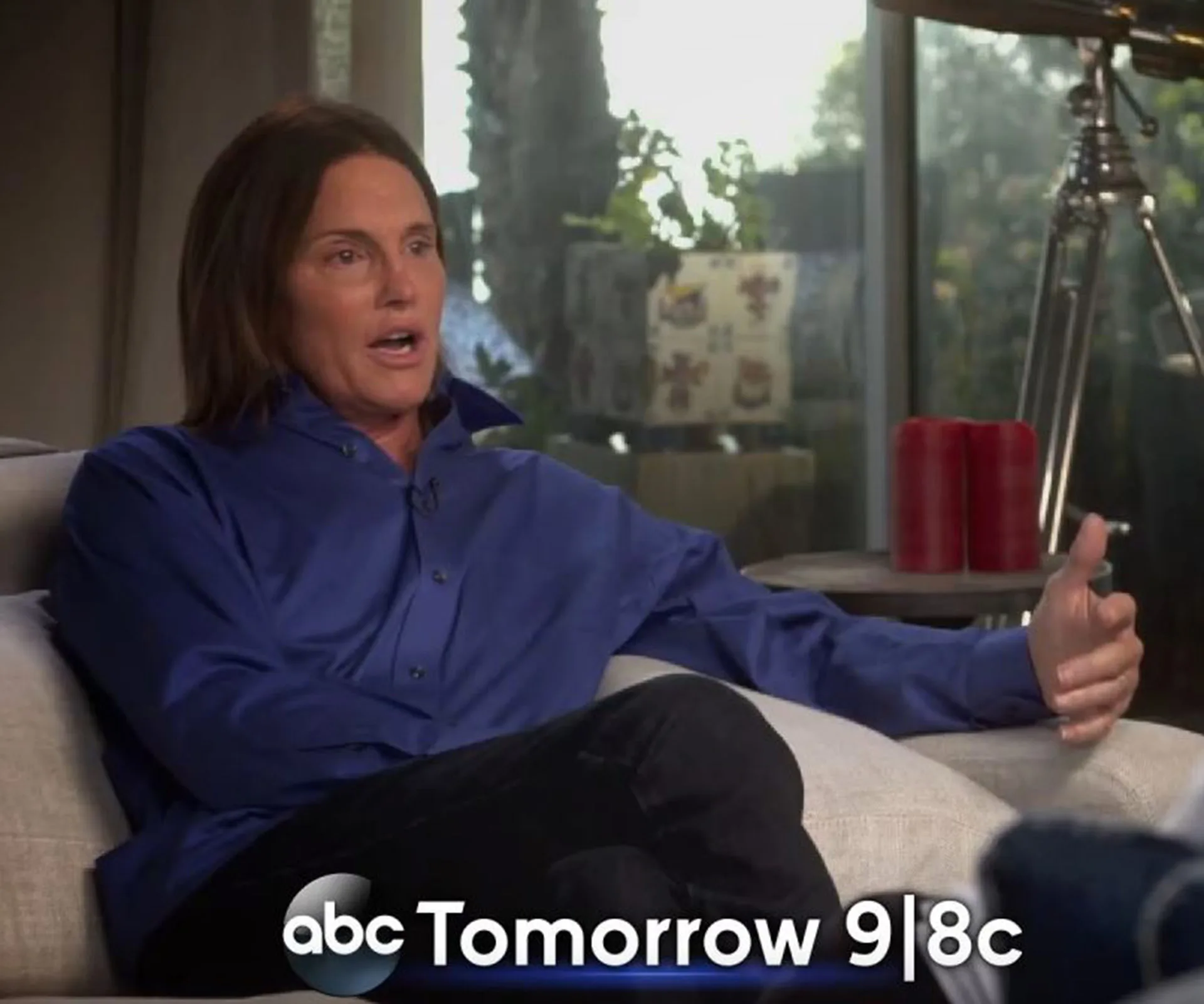 Person seated on a couch, wearing a blue shirt, gesturing while talking. ABC promo text at the bottom reads "Tomorrow 9|8c."