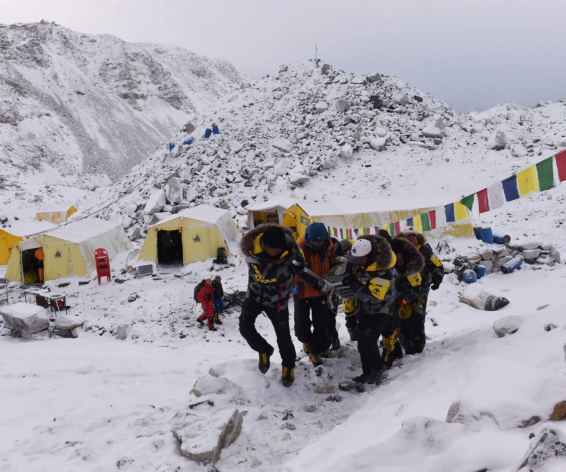 Rescuers carry a person on a stretcher through snow in an Everest Base Camp with tents and prayer flags visible.