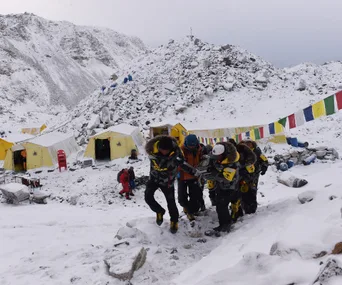 Rescuers carry a person on a stretcher through snow in an Everest Base Camp with tents and prayer flags visible.