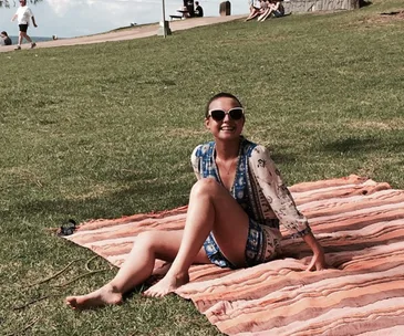A person sitting on a striped blanket in a park, smiling and wearing sunglasses and a patterned blouse.
