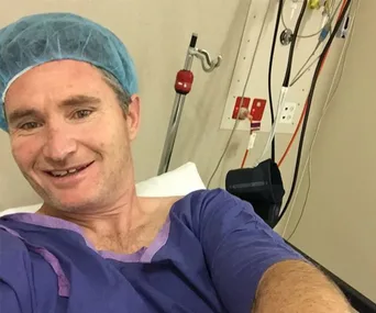 Man in hospital gown and cap takes selfie from hospital bed, smiling.