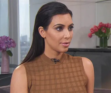 Kim Kardashian in a brown top, seated indoors with flowers in the background.