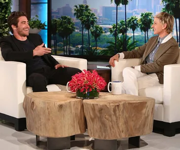"Two people sitting and talking on a talk show set with a scenic city backdrop and a wooden table with flowers."