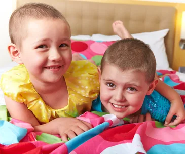 Two smiling young girls with short hair, lying on a colorful bed, wearing bright clothes.