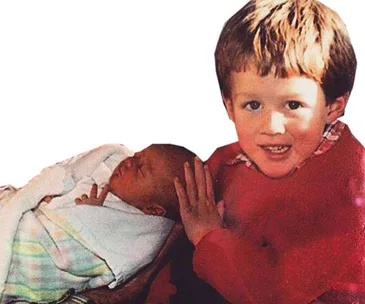 Two young children; a toddler smiling and gently touching a sleeping newborn wrapped in a pastel-patterned blanket.