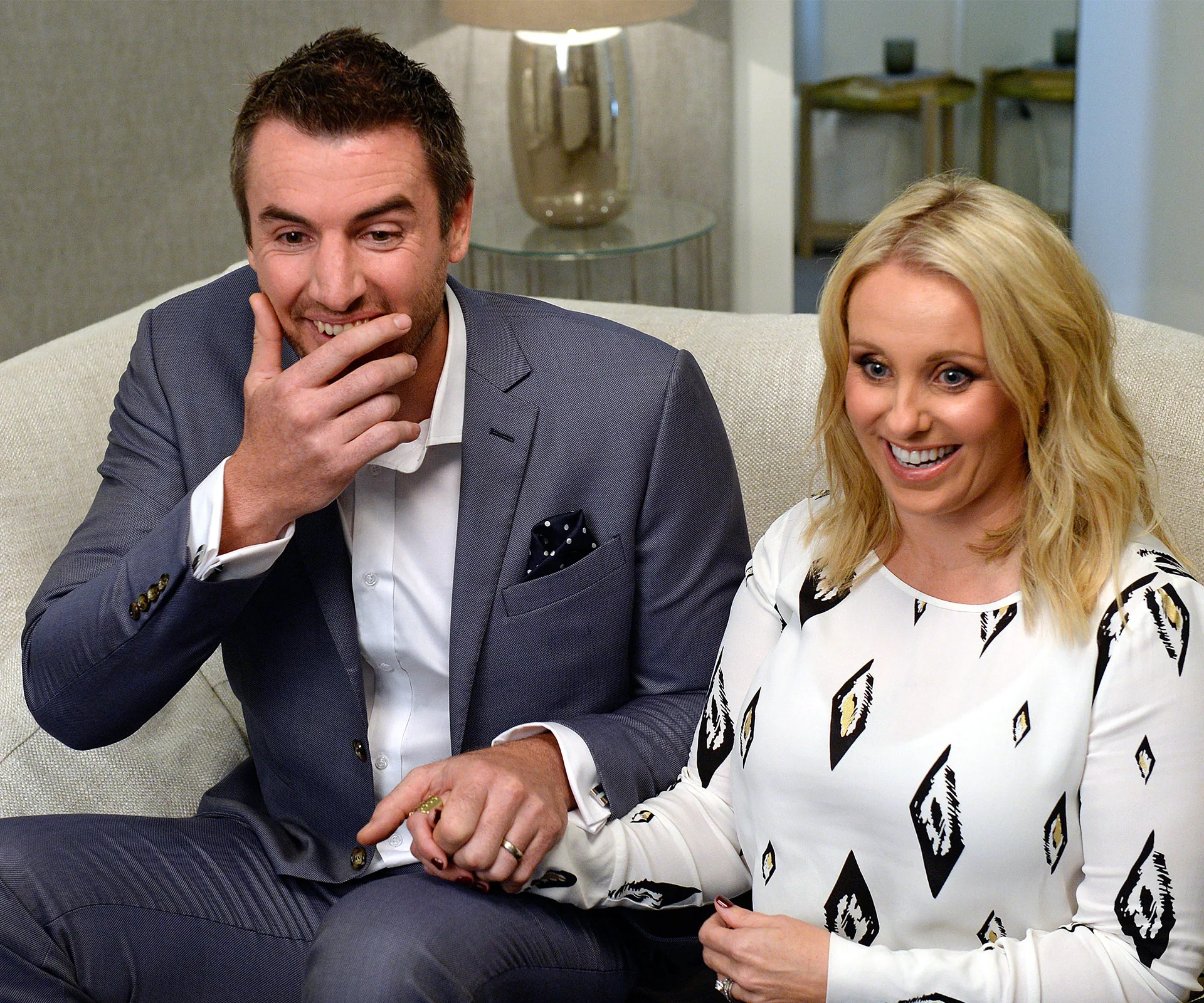 A man in a suit and a woman in a patterned dress sit on a couch, holding hands and smiling.