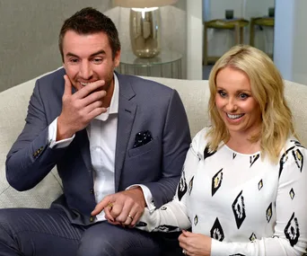 A man in a suit and a woman in a patterned dress sit on a couch, holding hands and smiling.