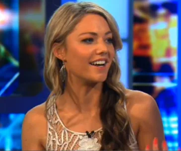 A woman with long hair and earrings, smiling on a TV show set with colorful blurred background.