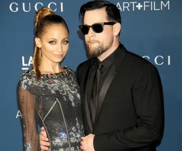 A couple posing at an event; woman in a sequined dress, man in a suit with sunglasses, blue backdrop with logos.