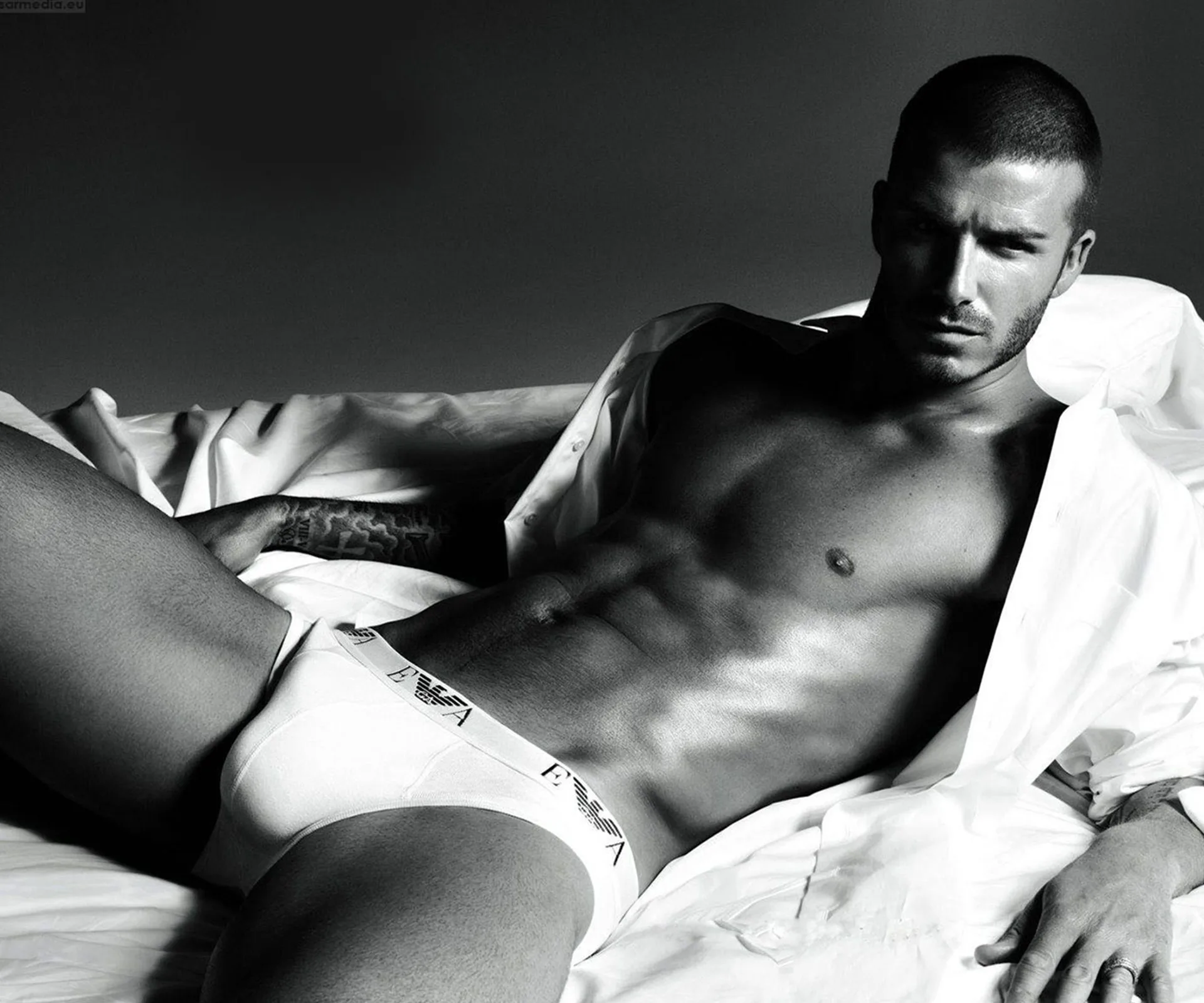 "David Beckham poses shirtless in white briefs on a bed, wearing a partially open white shirt. Black and white photo."