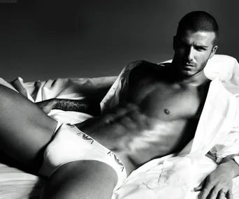 "David Beckham poses shirtless in white briefs on a bed, wearing a partially open white shirt. Black and white photo."