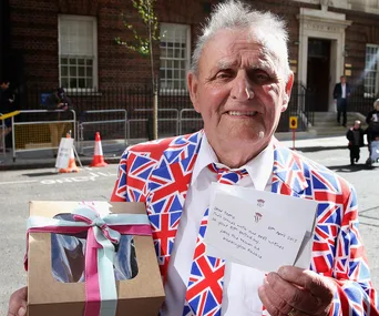 Man in a Union Jack suit holds a gift box and a handwritten note outside a brick building.