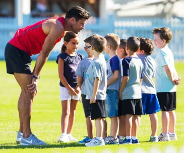 Man in athletic wear bends down, smiling and talking to a group of children standing on a grassy field.