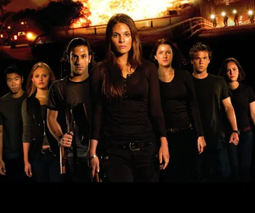 Group of young adults in dark clothing, some holding weapons, walking intensely with a fiery explosion in the background.