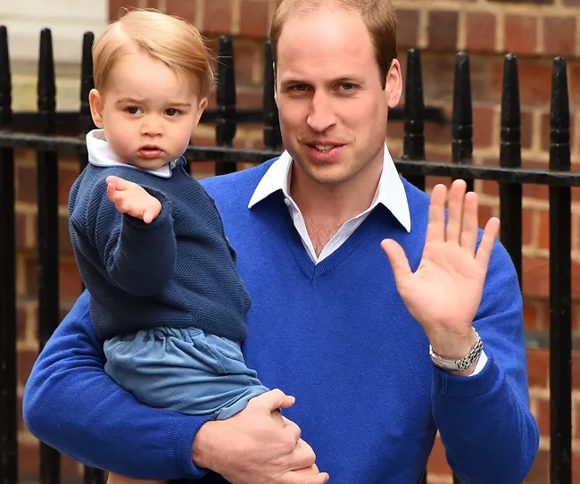 Father and young child wearing blue sweaters, waving towards photographers outside a brick building.