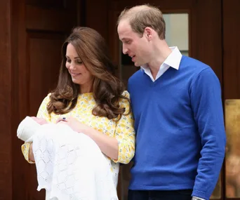 Royal couple holds newborn wrapped in a white blanket, smiling outside a building.