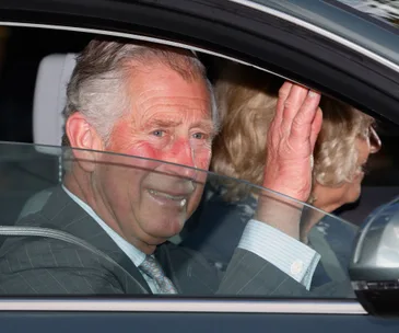 "Man in suit waves from car window with a woman sitting beside him, both smiling."