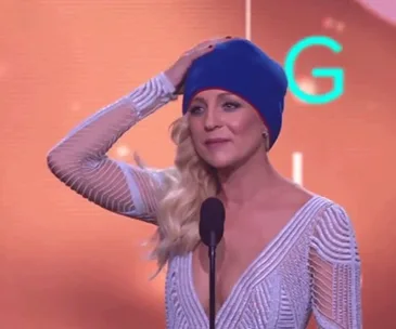 Carrie Bickmore wearing a blue beanie, holds a Logie award on stage, honoring her late husband.