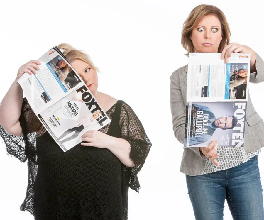 Two women with surprised expressions, holding Foxtel magazines, pose humorously against a white background.