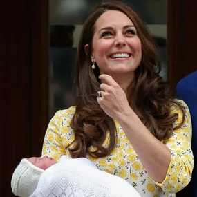 A smiling woman holding a newborn baby wrapped in a white blanket; she wears a yellow floral dress.