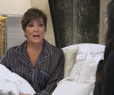 Kris Jenner in striped pajamas, appearing emotional, sits on a bed during a conversation.