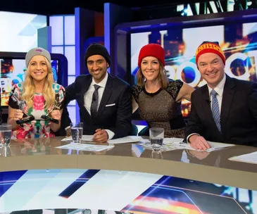 Four TV hosts wearing various beanies sit at a desk with awards, smiling at the camera.