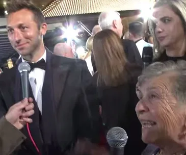 A man in a tuxedo smiles at an event, while multiple people hold microphones in a crowded, well-lit room.