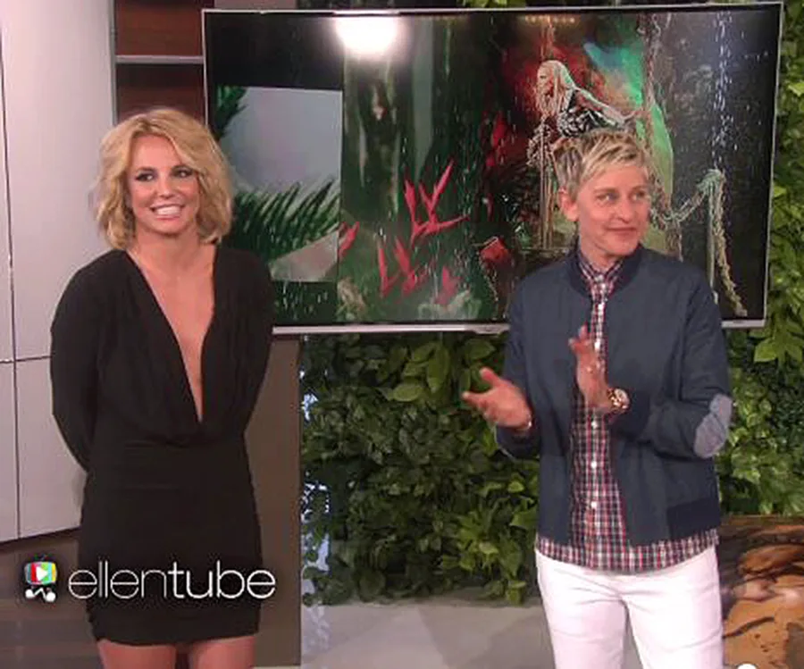 Two people smiling on a talk show set with a screen displaying colorful graphics behind them.