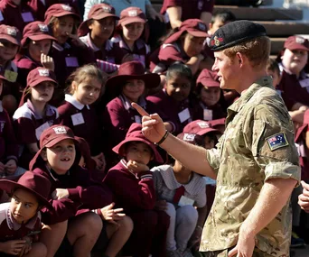 Prince in uniform speaking to schoolchildren wearing maroon uniforms and hats, engaging group outdoors.