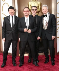 Members of U2 pose on the red carpet in suits in front of an Oscar statue.
