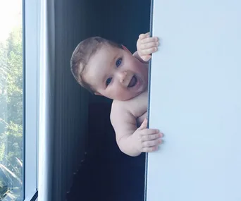 A baby peeks playfully around a white door, smiling with wide eyes, next to a window with trees visible outside.