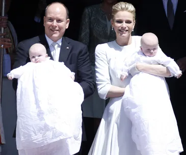 A couple in formal attire holding twin babies in white christening gowns outdoors.