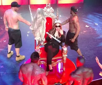 Mariah Carey sits on a throne holding a scepter, surrounded by shirtless dancers on stage.
