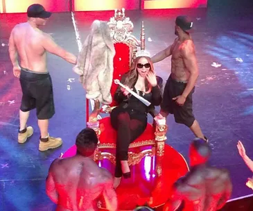 Mariah Carey sits on a throne holding a scepter, surrounded by shirtless dancers on stage.