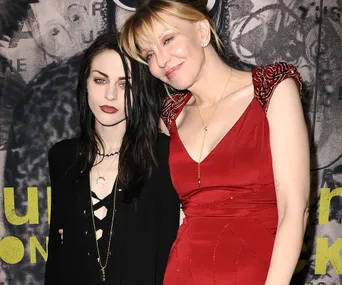 Two people posing together, one in a black outfit and the other in a red dress, with a patterned background.